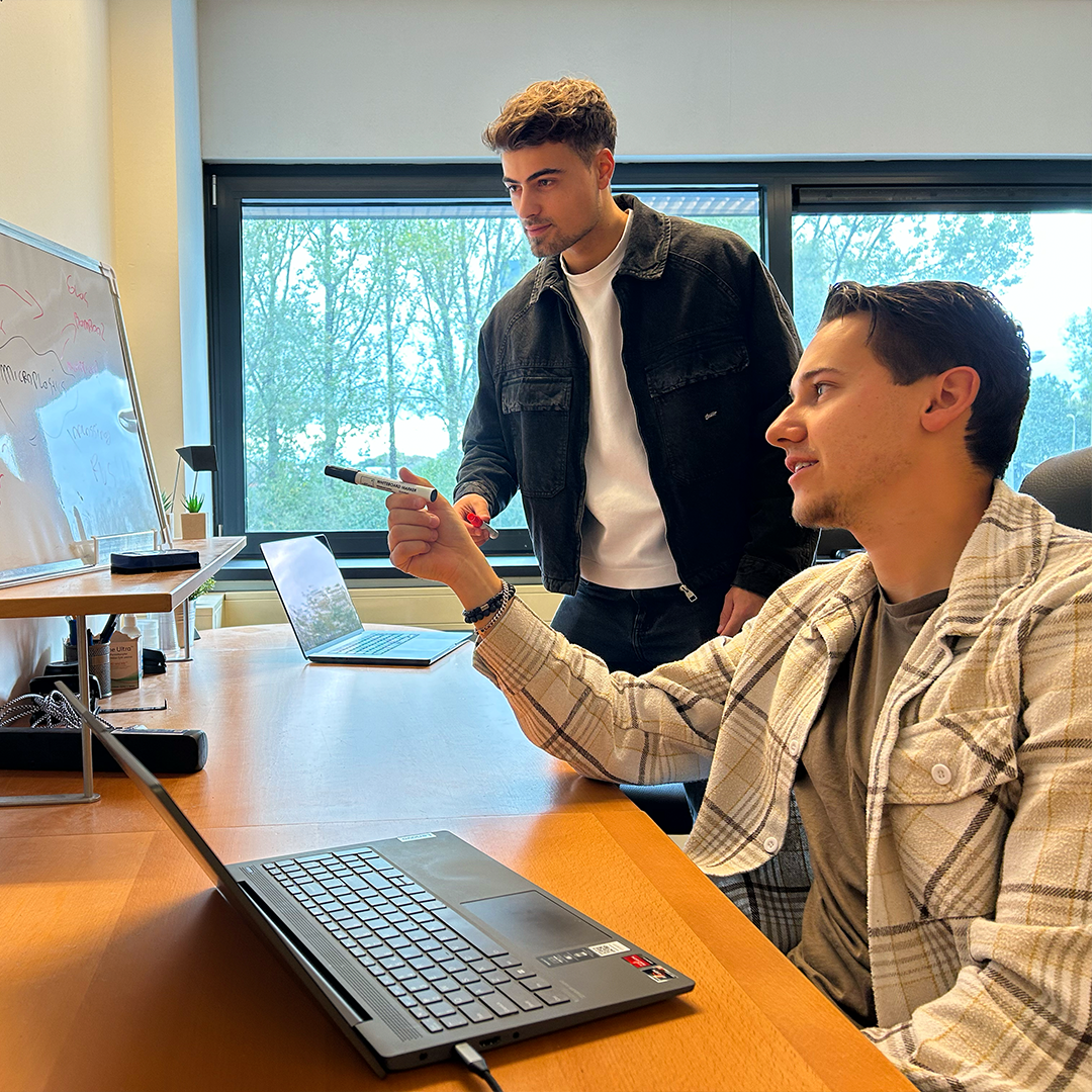 Twee collega’s die samenwerken aan een project in een kantooromgeving, één wijst iets aan op een laptop.